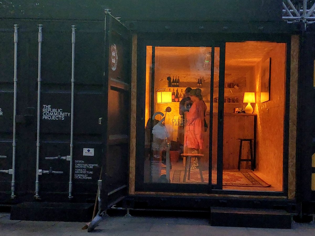 The taproom's glass front open at dusk, warm light inside and steps up into the container
