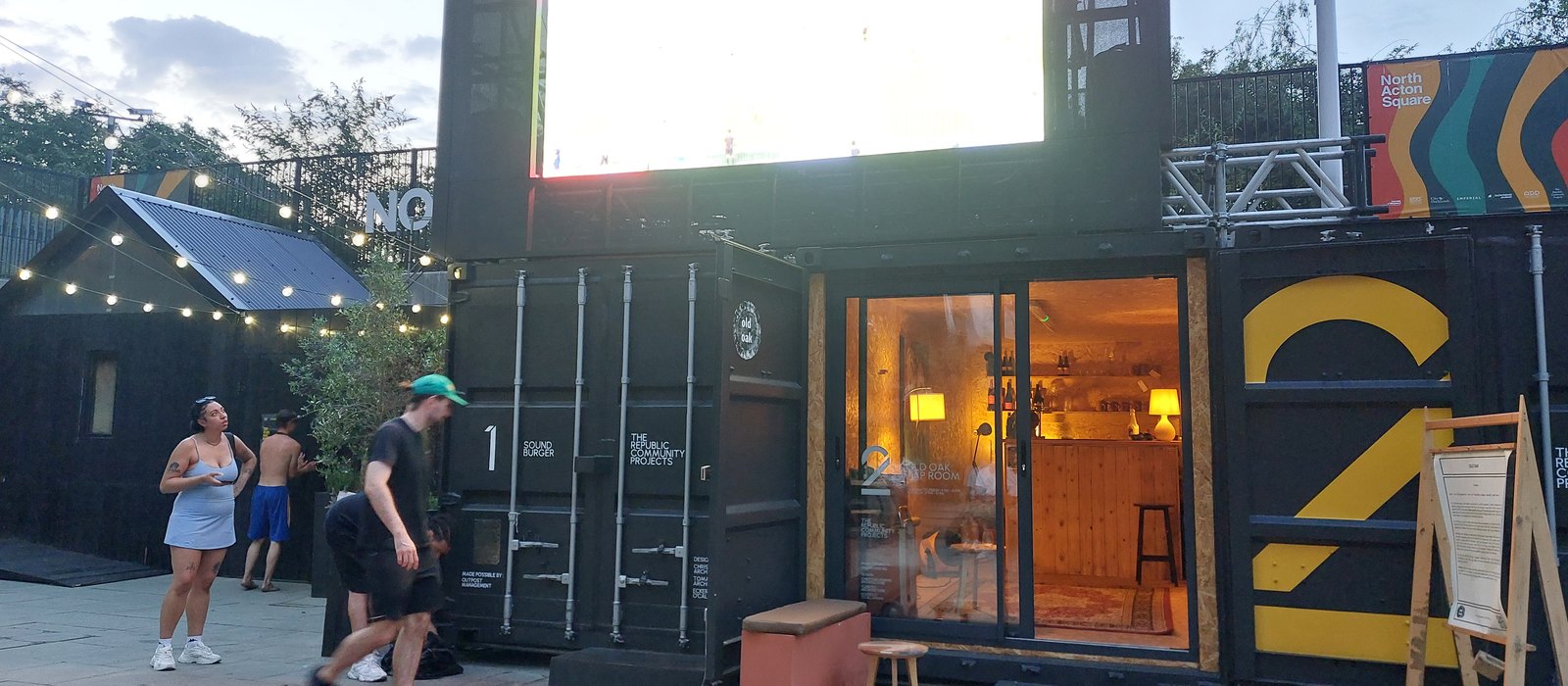 The Old Oak Tap Room lit up in container 2 on North Acton Square in the evening, with festoon lights and a big screen above the units